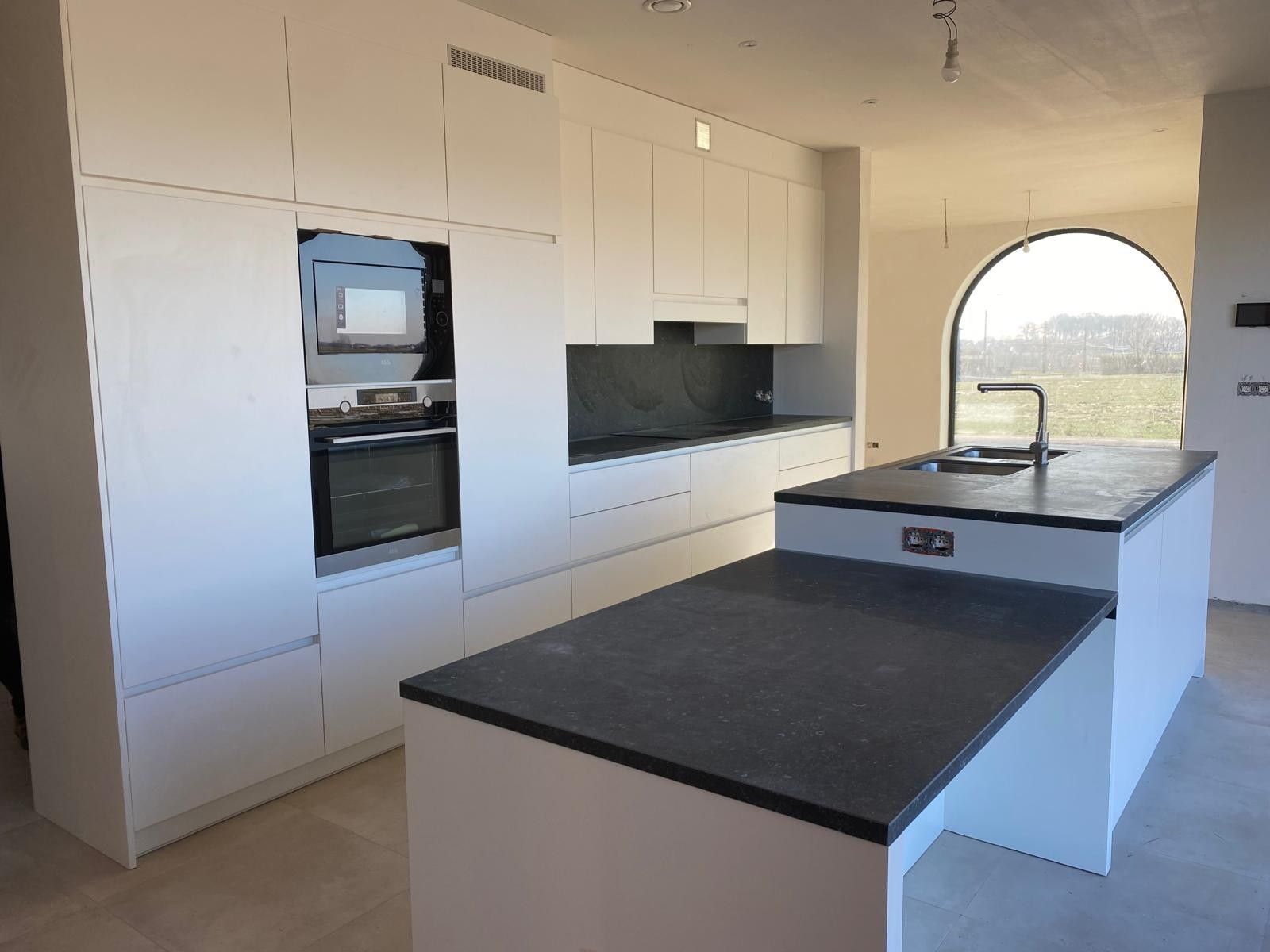 Witte greeploze keuken met een oven en een microgolf in een hoge kast, een donkere achterwand en een eiland op twee hoogtes voor een boogvenster