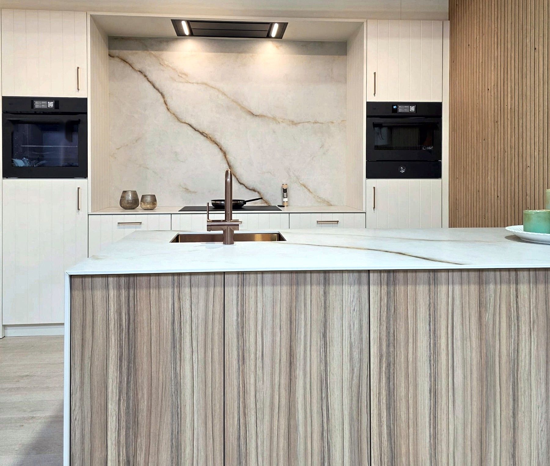 Keuken met crèmekleurige fronten, een achterwand in marmerlook en een eiland met houten fronten en een koperkleurige kraan