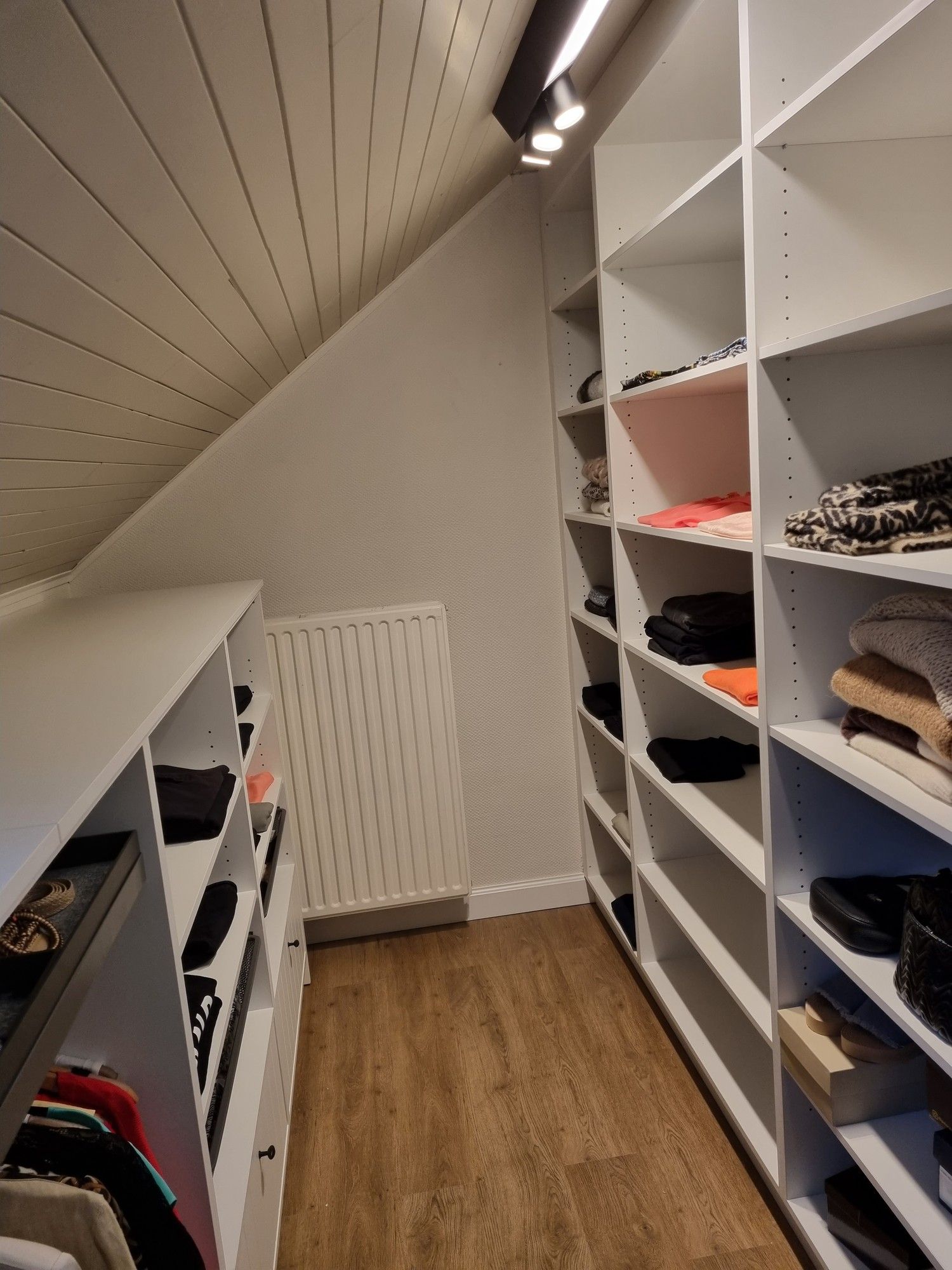 Dressing onder een schuin dak met witte open kasten aan beide kanten en spots op een rail