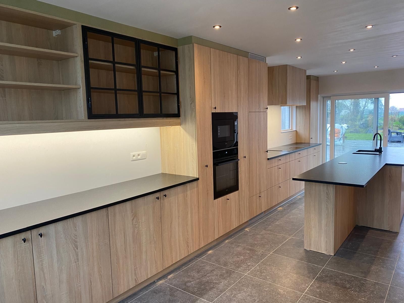 Lange keukenwand in een houttint met open planken, een glaskast met zwart kader, een oven en een microgolf en een eiland met een donker werkblad