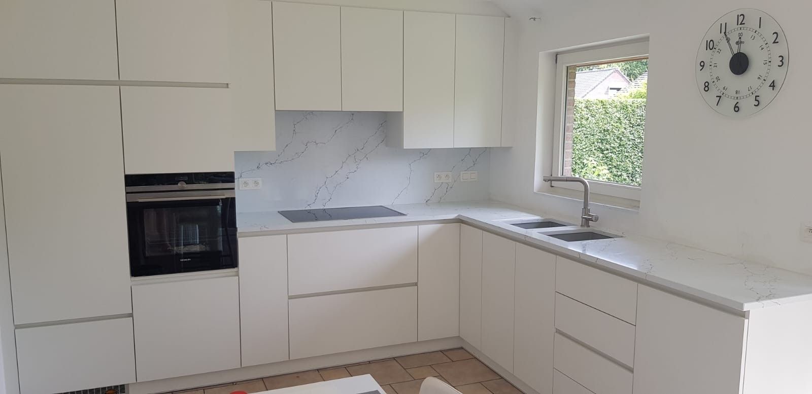 Na: een witte greeploze keuken in hoekopstelling met een werkblad en achterwand in marmerlook en een inbouwoven