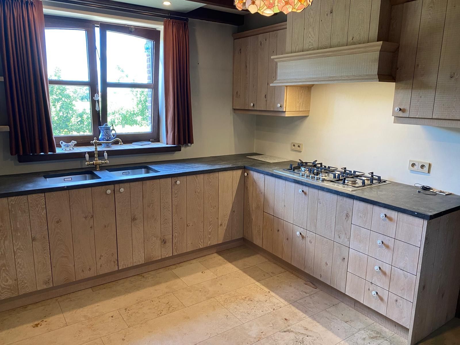 Landelijke keuken in hoekopstelling met fronten van verticale planken, een dubbele spoelbak onder het raam en een gaskookplaat onder een schouw