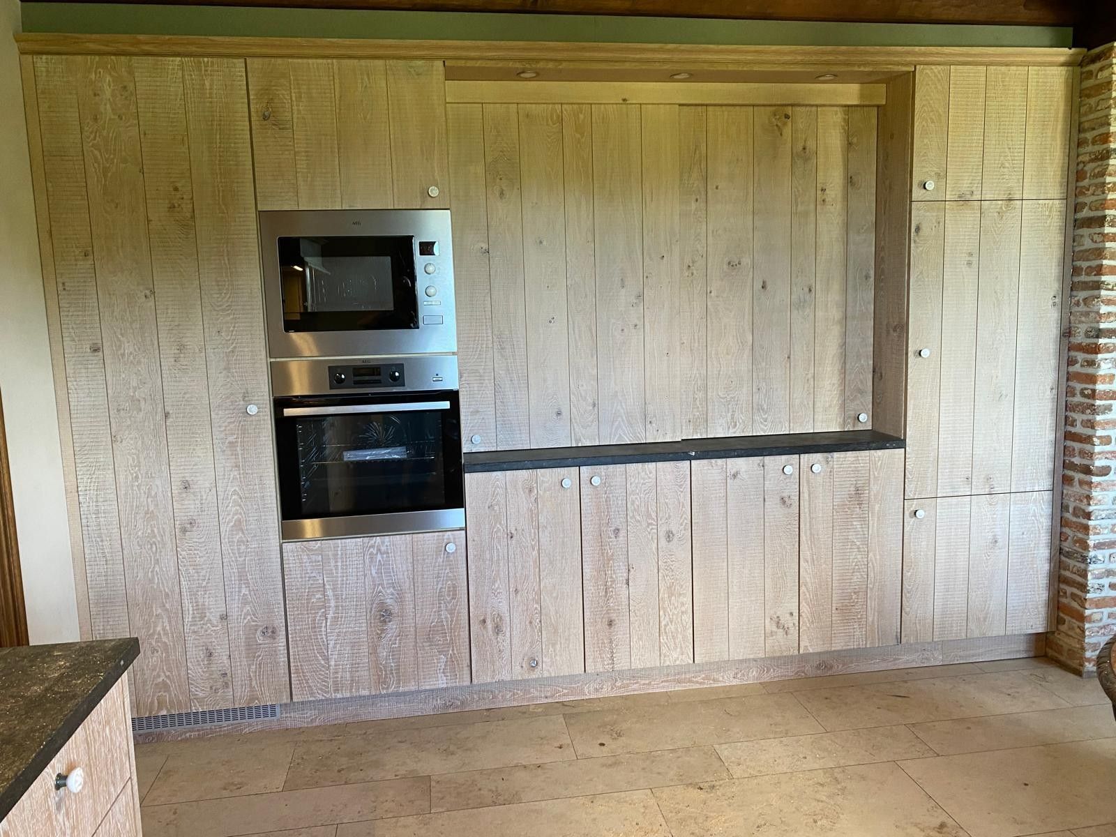 Hoge kastenwand met fronten van planken, een oven en een microgolf en een nis met een werkblad, naast een bakstenen muur