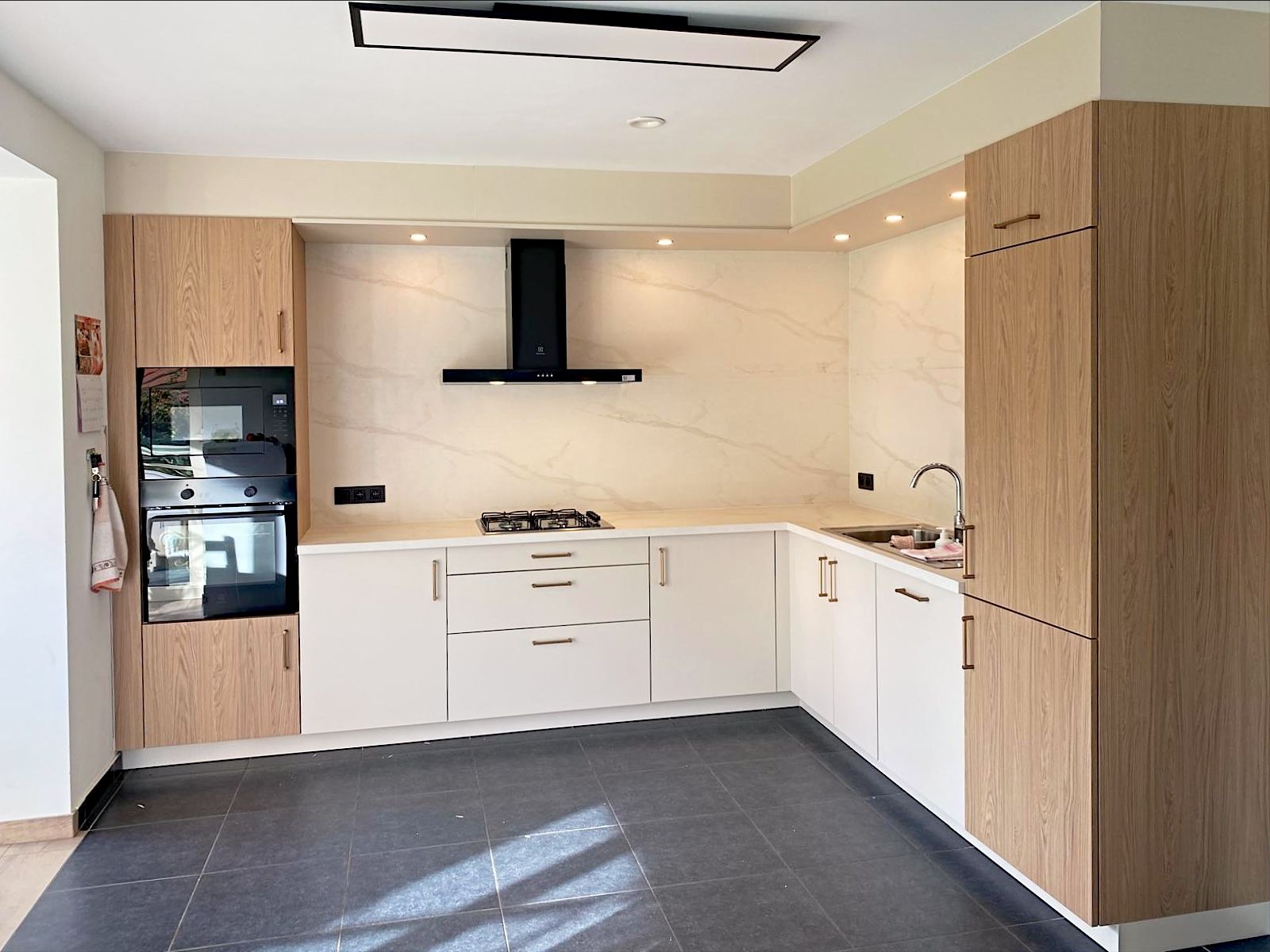 L-vormige keuken met witte onderkasten, hoge kasten in een warme houttint en een achterwand in marmerlook